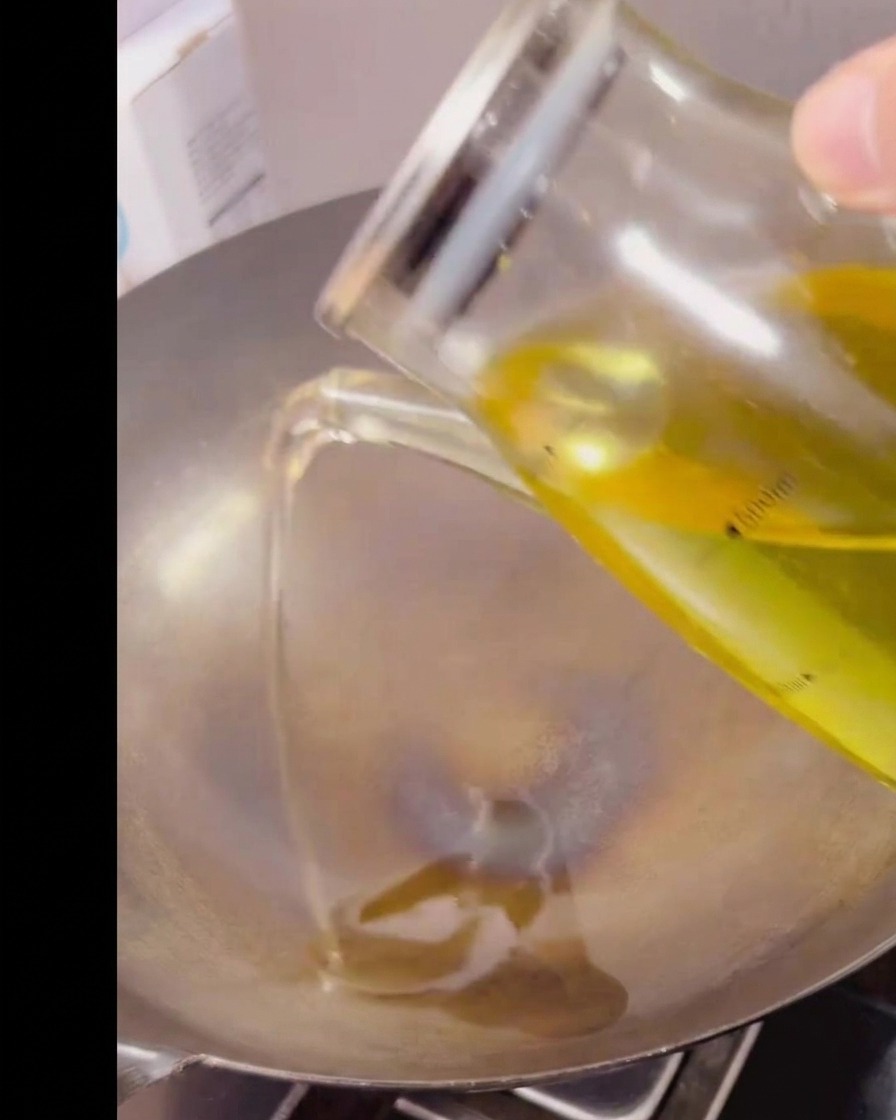 Klares Speiseöl wird aus einem Messbecher aus Glas in einen heißen, leeren Metallwok gegossen.