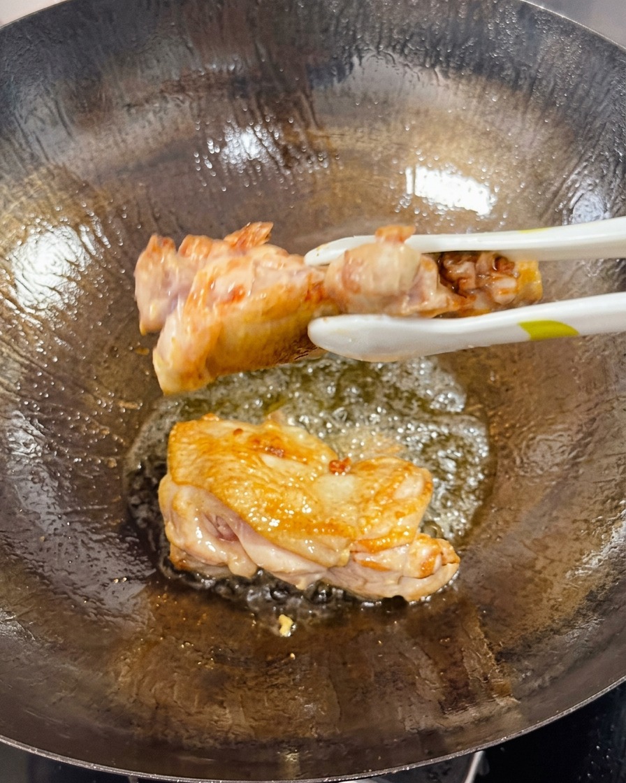 Eine Zange wendet eine angebratene Hähnchenoberkeule im Wok und zeigt die knusprige, goldbraune Seite.