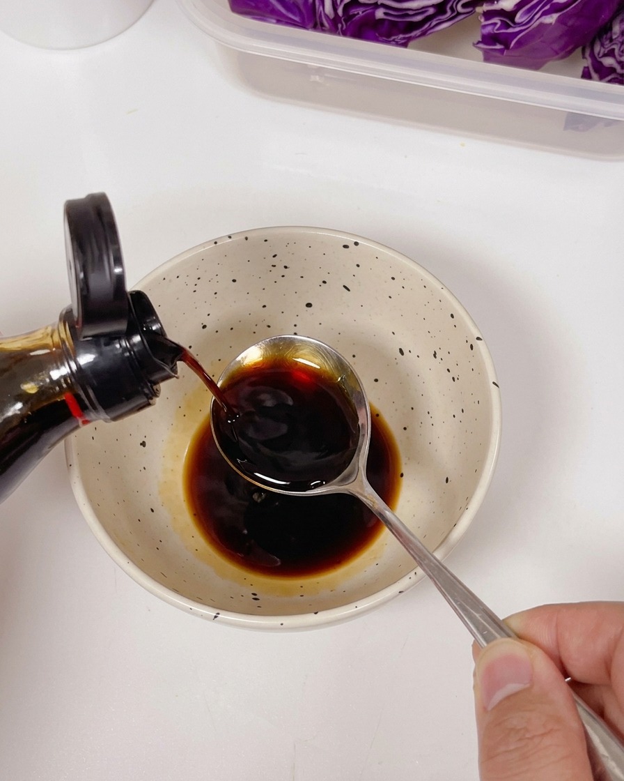 Helle Sojasauce wird aus einer Flasche auf einen Löffel über einer Schüssel mit dunkler Sauce und Gewürzen gegossen.