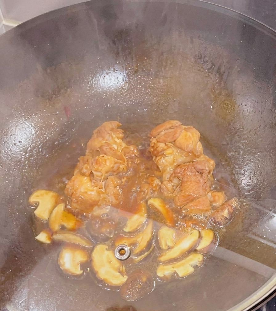 Hähnchenschenkel und geschnittene Shiitake-Pilze köcheln in einer dunklen Sauce in einem Wok unter einem Glasdeckel.