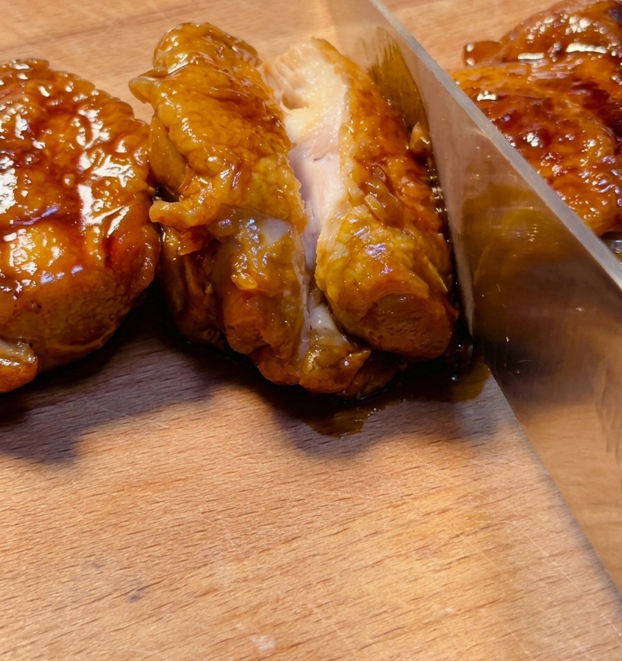 Ein scharfes Messer schneidet eine tief glasierte Teriyaki-Hähnchenoberkeule auf einem Holzbrett auf und zeigt das saftige Fleisch im Inneren.