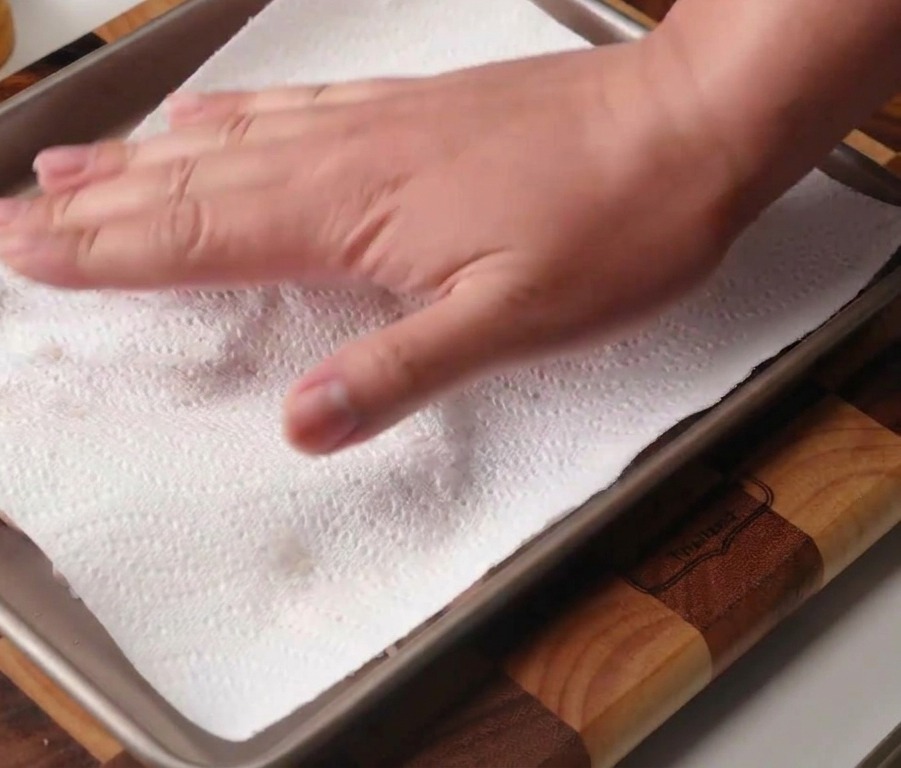 Eine Hand tupft rohe Hähnchenkeulen auf einem Backblech mit einem Küchentuch trocken.
