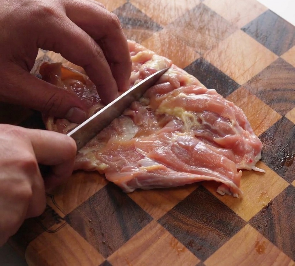 Eine Hand klopft ausgelöste Hähnchenkeulen mit dem Messerrücken auf einem Holzschneidebrett.