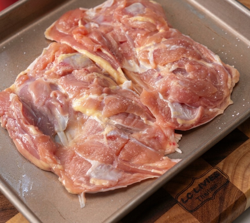 Rohe, ausgelöste Hähnchenkeulen auf einem Blech, bestreut mit gehacktem Knoblauch und schwarzem Pfeffer.