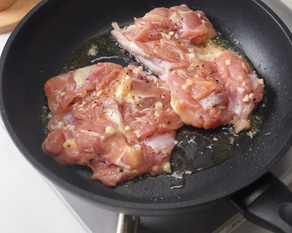 Zwei marinierte Hähnchenkeulen werden mit der Hautseite nach unten in einer dunklen, beschichteten Pfanne angebraten.