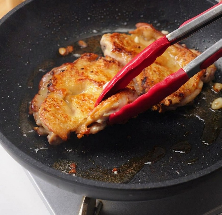 Eine Hand gießt dunkle Teriyaki-Sauce aus einer Glasschüssel über die angebratenen Hähnchenkeulen in der Pfanne.