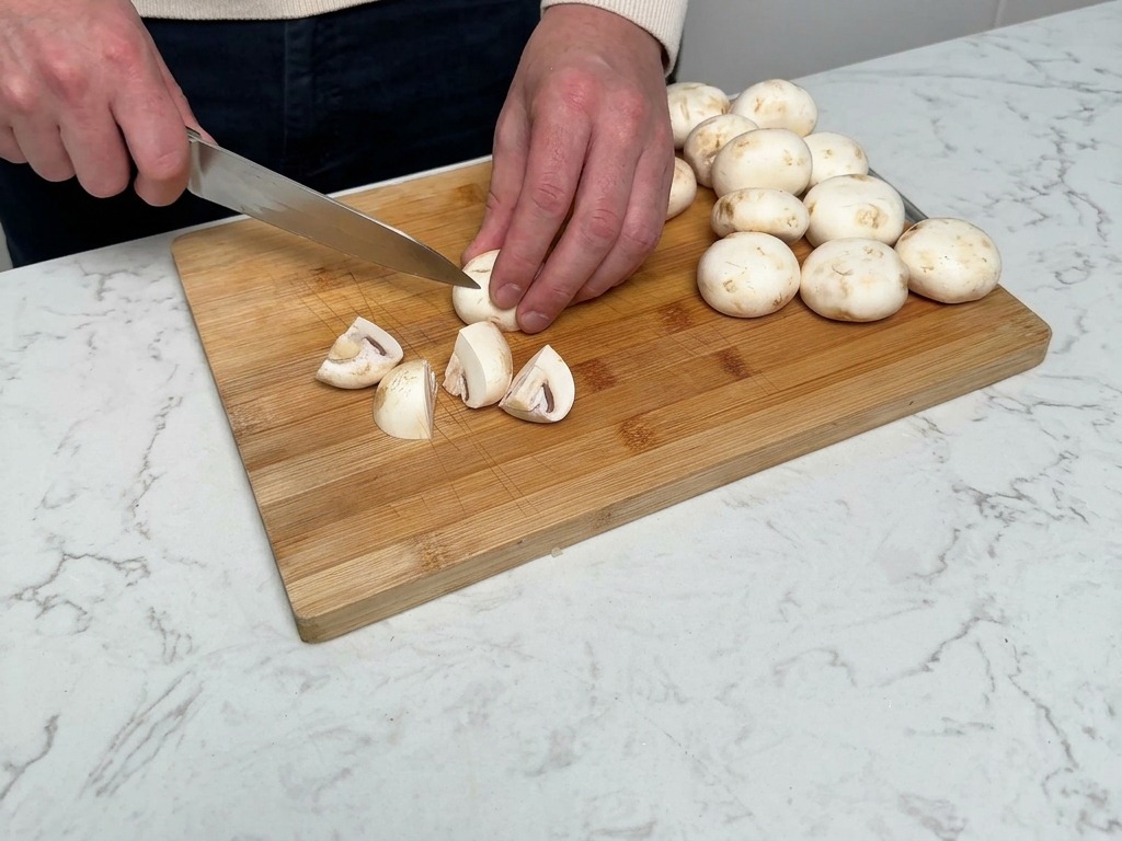 Champignons werden auf einem Holzbrett mit einem Kochmesser gevierteelt.