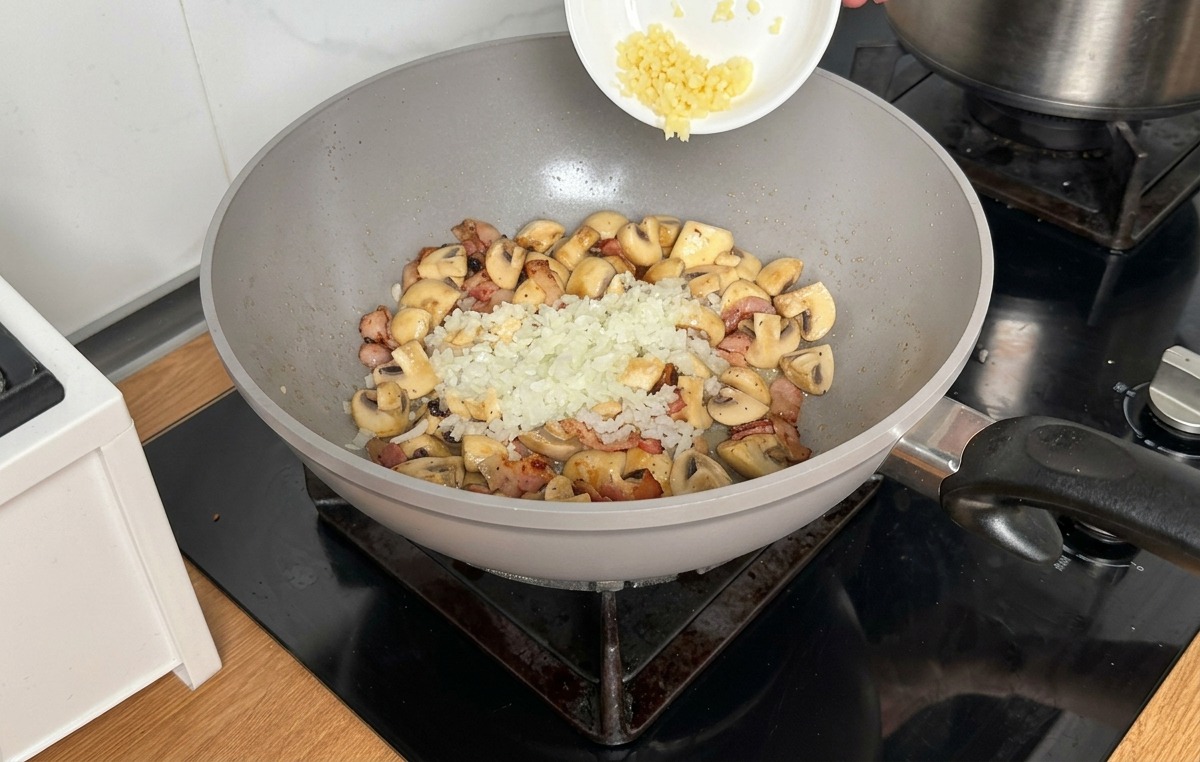 Fein gewürfelte Zwiebeln und Knoblauch werden über die Pilze und den Speck in die Pfanne gestreut.