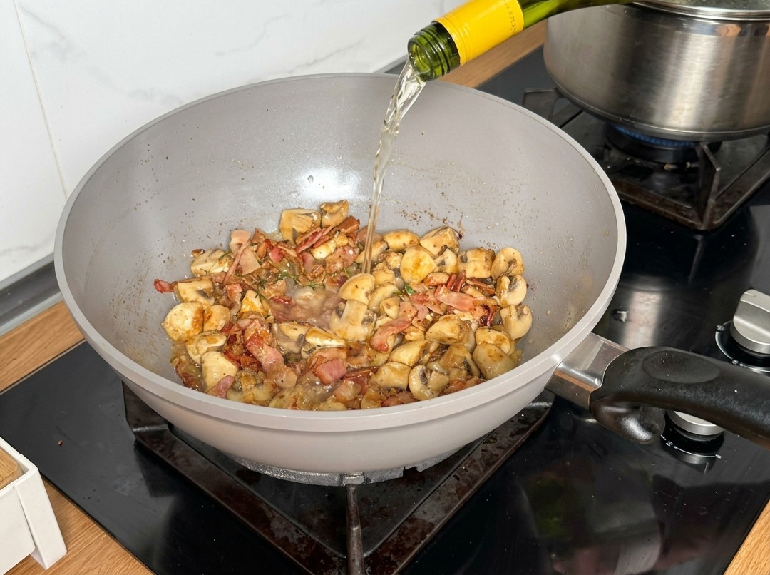 Weißwein wird aus einer Flasche in die Pfanne zu den Pilzen gegossen.