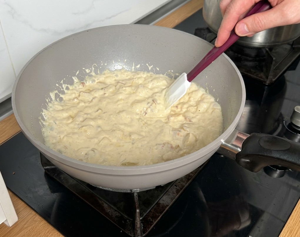 Ein Teigschaber rührt in der blubbernden Sahnesauce mit Speck und Pilzen.