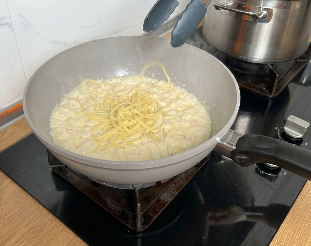 Eine Küchenzange hebt die fertige Pasta direkt aus dem Wasser in die Pfanne.