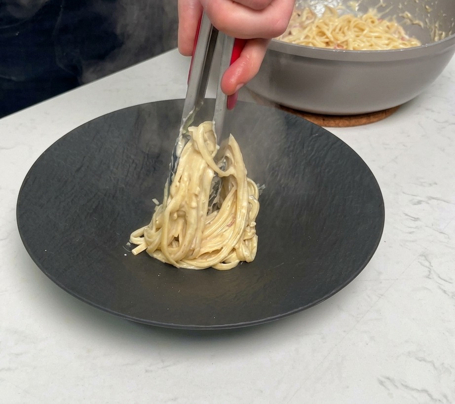 Mit einer Zange wird eine Portion cremige Pasta auf einen schwarzen Teller gedreht.