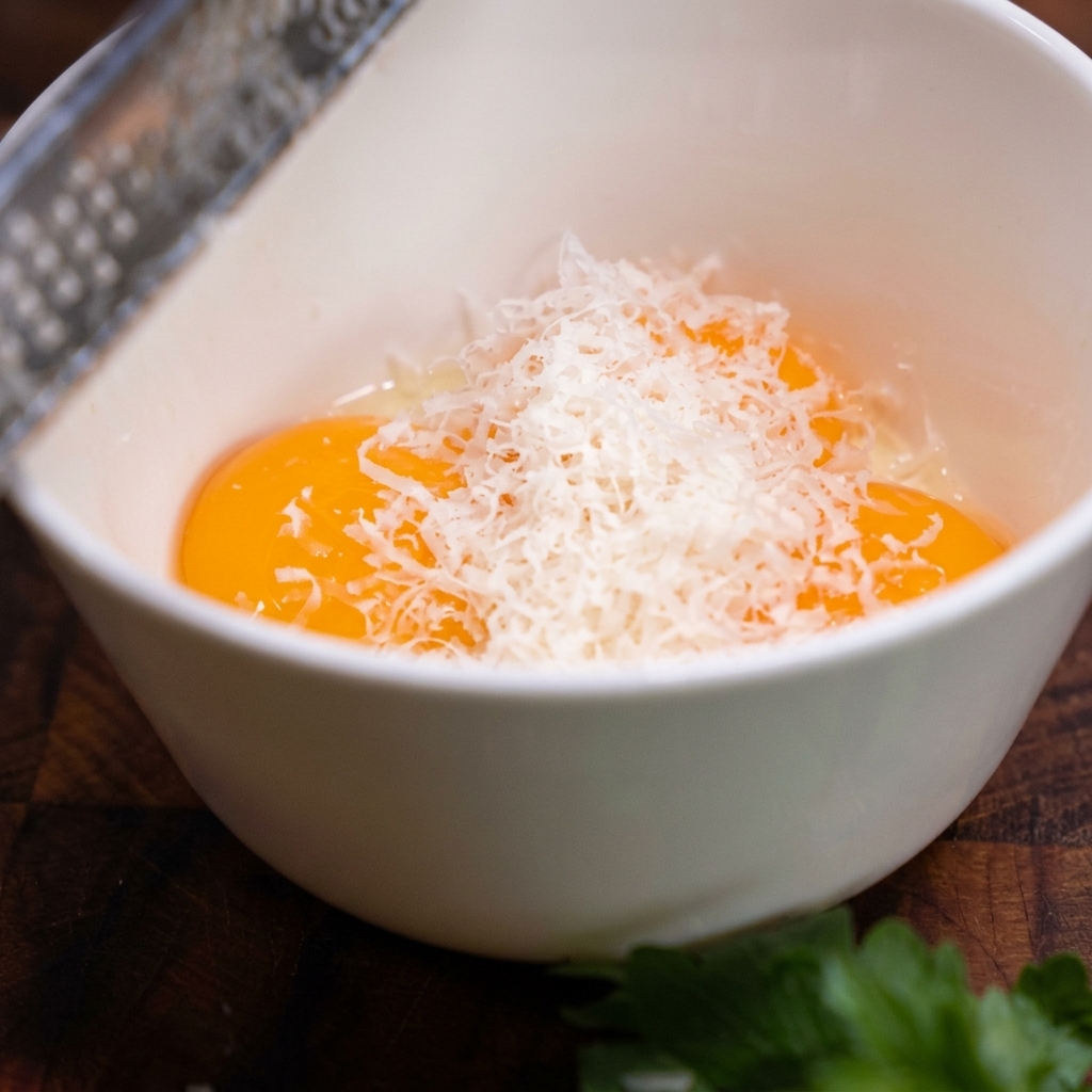 Eine Metallreibe hobelt feine weiße Parmesanspäne in eine Schüssel mit drei Eigelben.