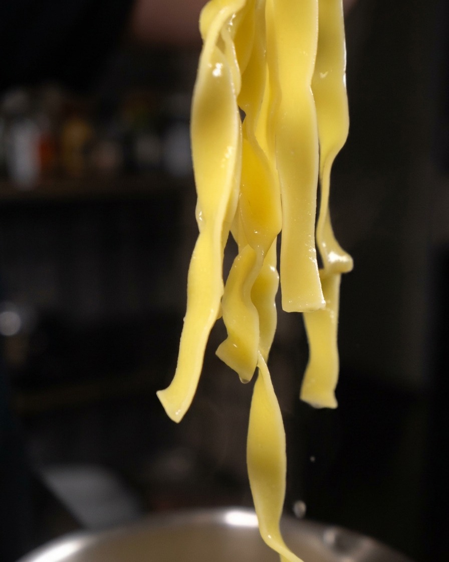 Lange, gelbe Tagliatelle-Stränge hängen an einer Küchenzange und tropfen ab.