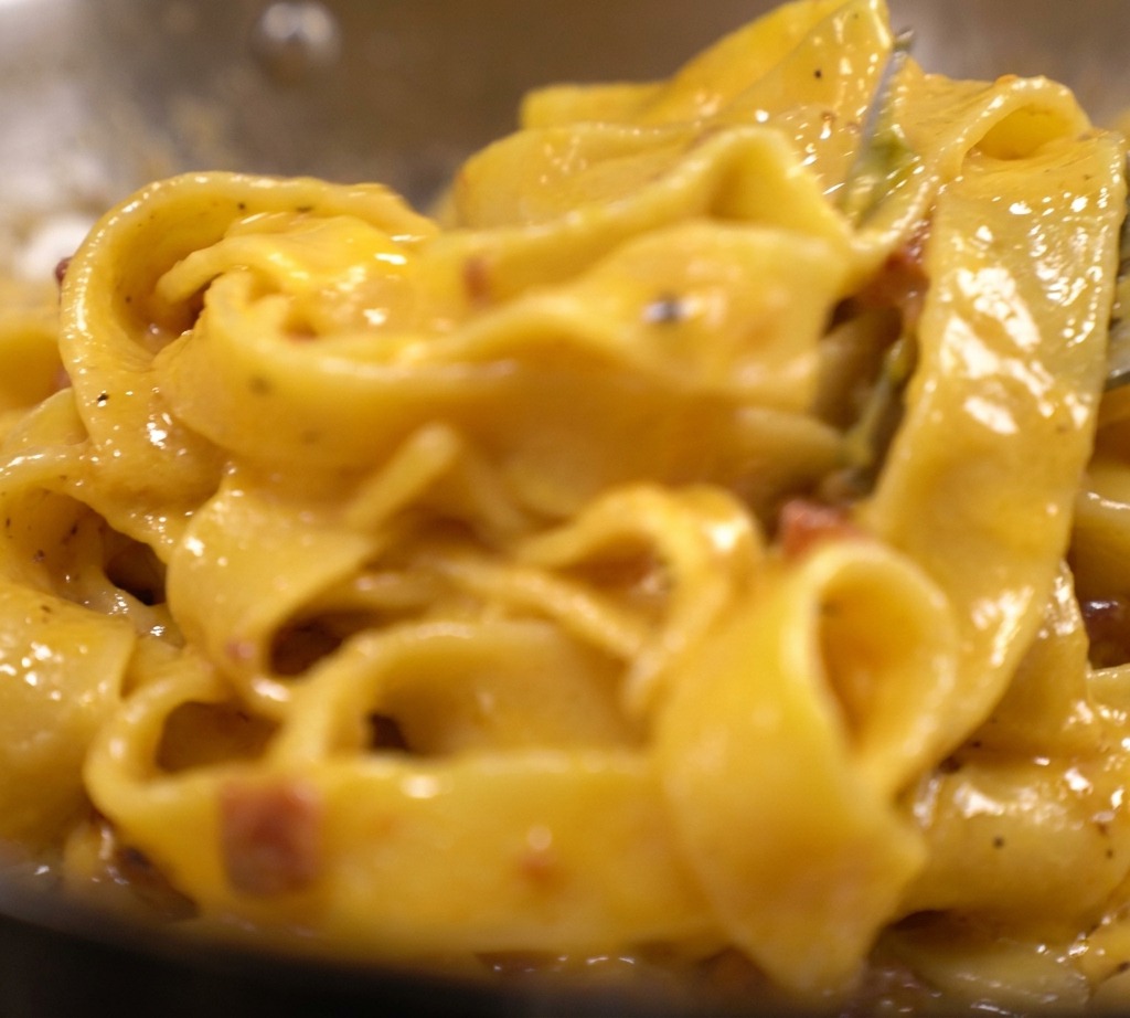 Tagliatelle in einer dicken, leuchtend gelben Ei-Käse-Sauce mit Speck in der Pfanne.
