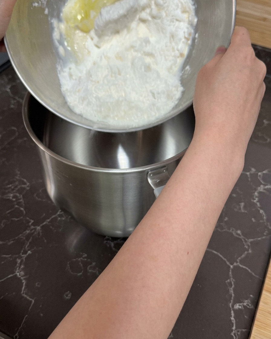 Hände gießen eine grobe Mischung aus Mehl und feuchten Zutaten in die Schüssel einer Küchenmaschine.