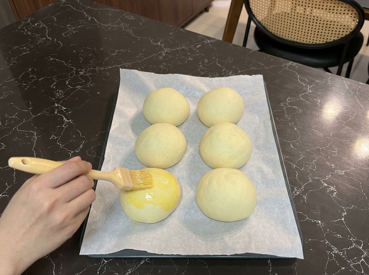 Eine Hand bestreicht mit einem Backpinsel die Oberseite eines aufgegangenen Burger-Buns mit einer glänzenden Eigelb-Mischung.