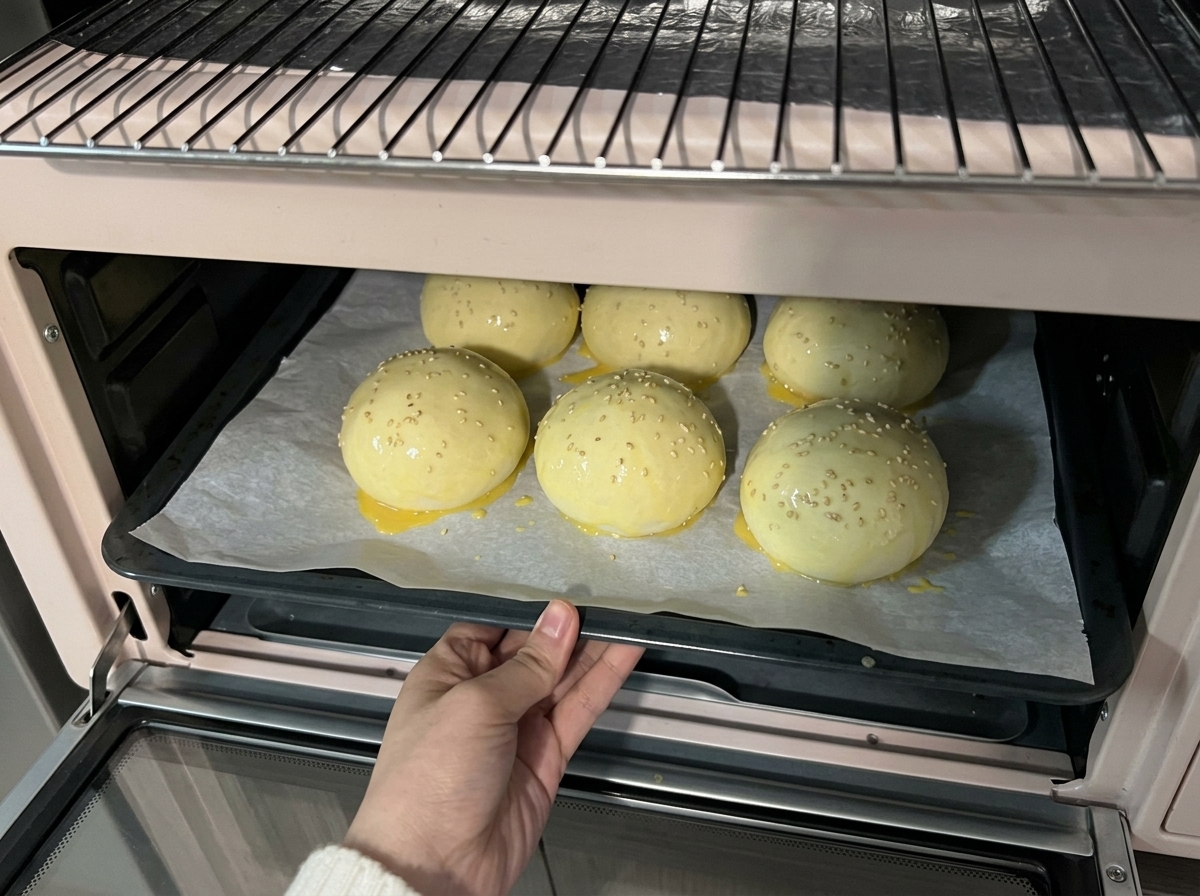 Ein Blech mit mit Sesam bestreuten Teigkugeln wird in einen hellrosa Minibackofen zum Backen geschoben.