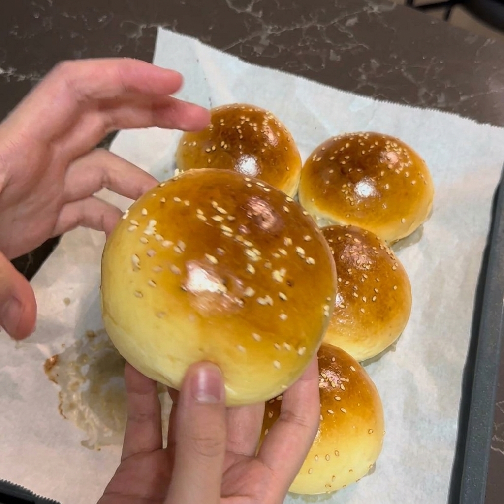 Hände halten ein frisch gebackenes, goldbraunes Burgerbrötchen mit weißem Sesam.