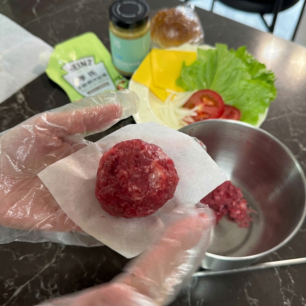 Eine behandschuhte Hand hält eine Kugel aus rohem Rinderhackfleisch auf einem kleinen Quadrat aus weißem Backpapier.