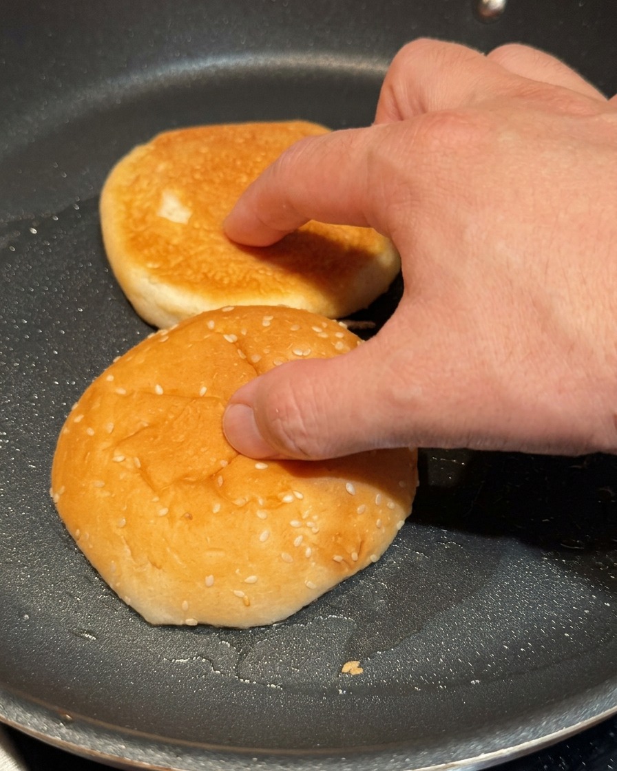 Eine Hand drückt zwei Hälften eines Burger-Brötchens zum Toasten in eine heiße Pfanne.