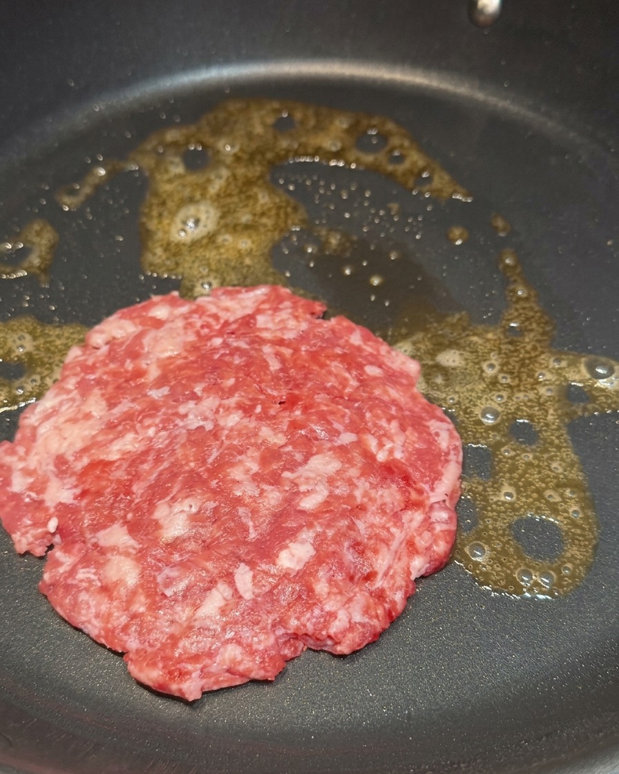 Ein rohes Rindfleisch-Patty brät zischend in geschmolzener Butter in einer Pfanne.