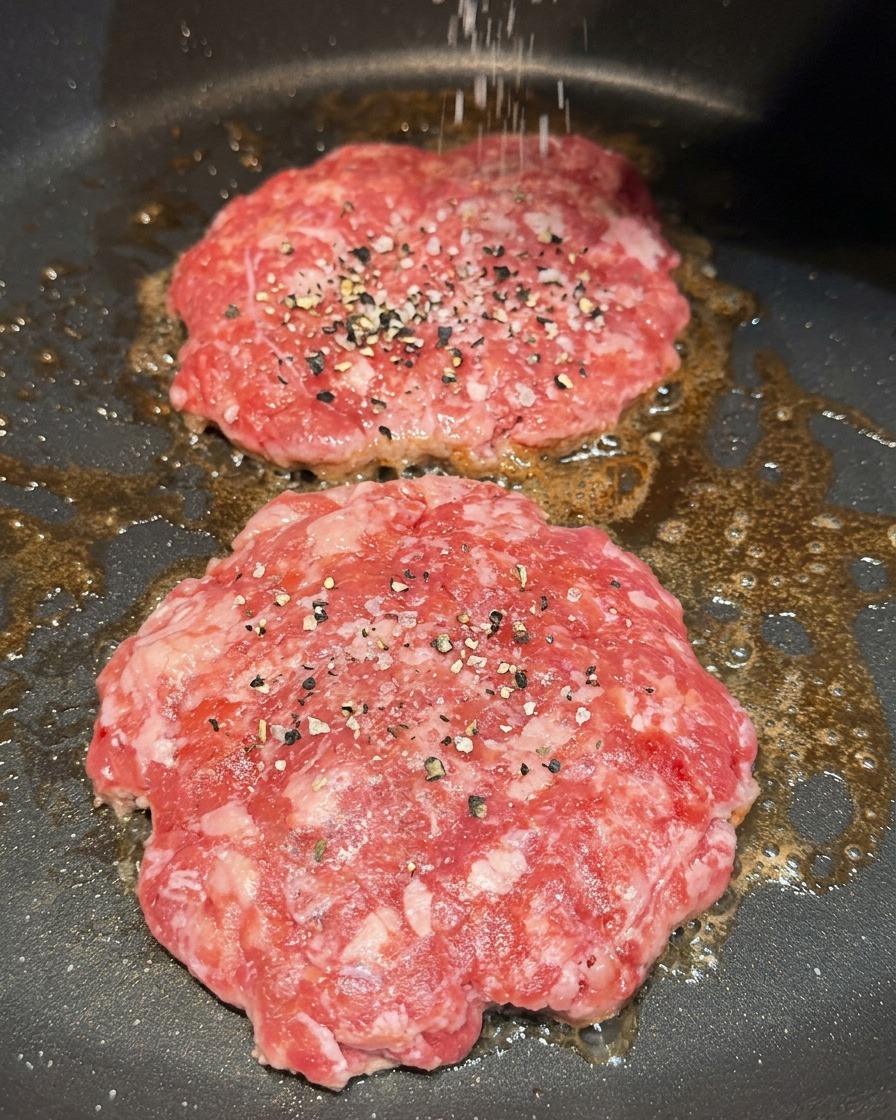 Grobes Salz und schwarzer Pfeffer werden über zwei bratende Fleisch-Patties gestreut.