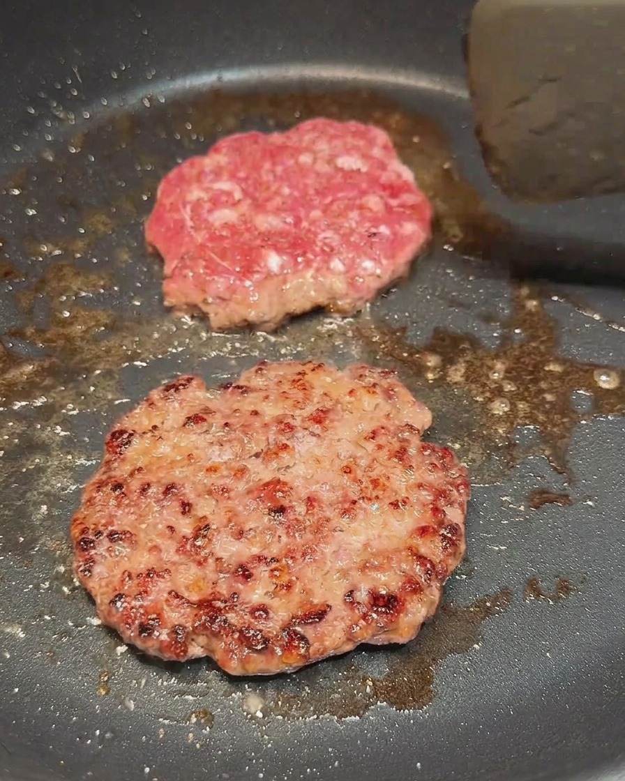 Zwei Fleisch-Patties in der Pfanne, eines davon wurde gerade gewendet und zeigt eine braune Kruste.