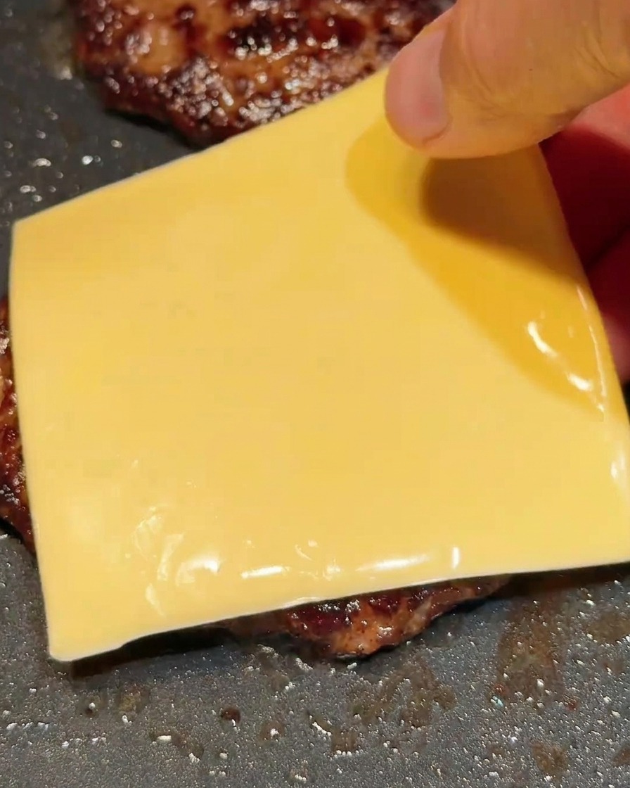 Eine Hand legt eine Scheibe gelben Käse auf ein heißes Fleisch-Patty in der Pfanne.