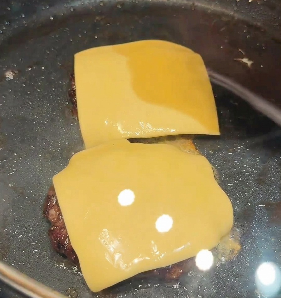 Zwei Käsescheiben schmelzen auf den Fleisch-Patties in einer abgedeckten Pfanne.