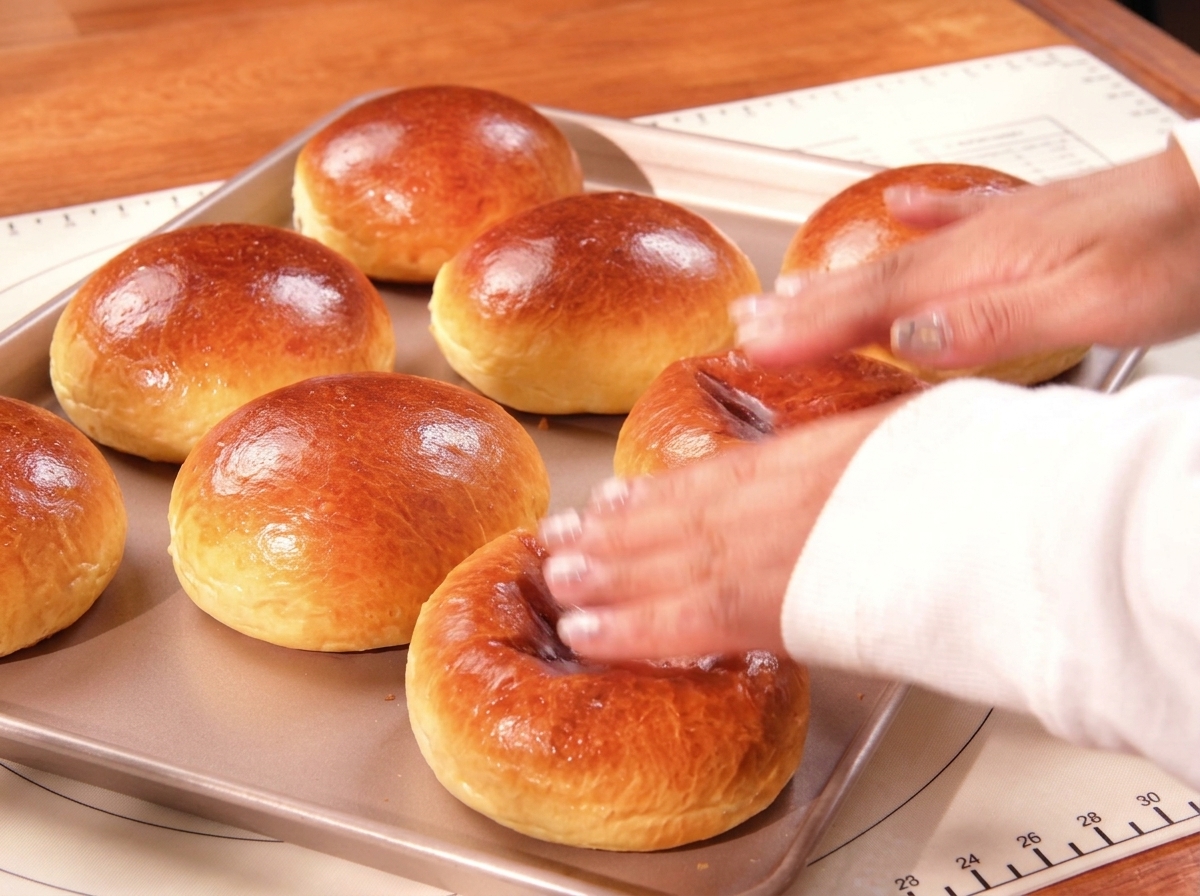 Eine Hand tippt vorsichtig auf frisch gebackene, goldbraun glänzende Brioche-Brötchen auf einem Backblech.