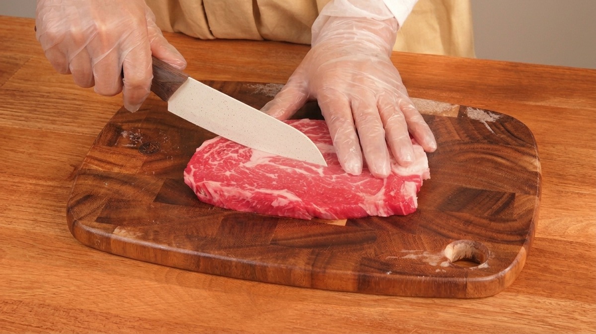 Hände schneiden ein dickes Stück marmoriertes Rindfleisch mit einem scharfen Messer auf einem Holzbrett.