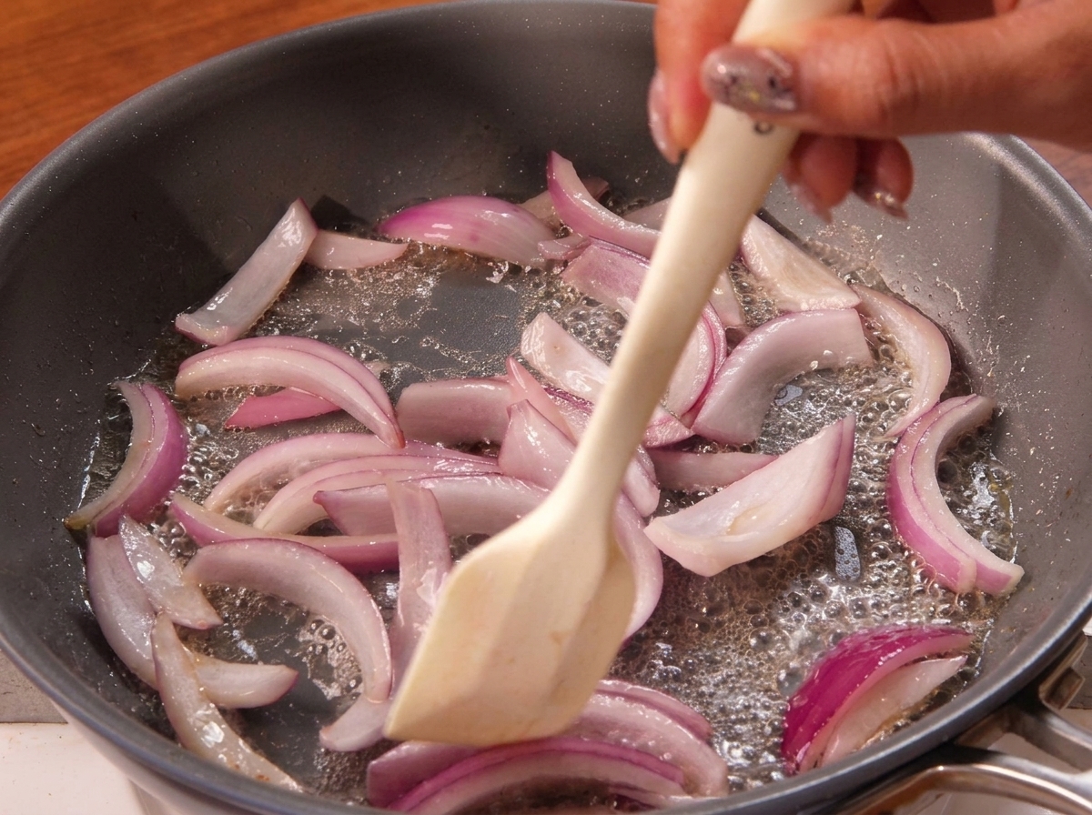 Ein Pfannenwender rührt rote Zwiebelstreifen in einer Pfanne um.