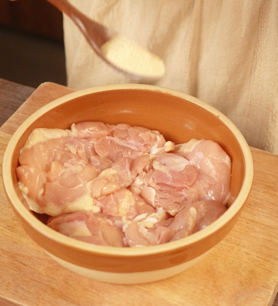 Ein Holzlöffel streut helles Knoblauchpulver über rohe, knochenlose Hähnchenoberschenkel in einer braunen Keramikschüssel.