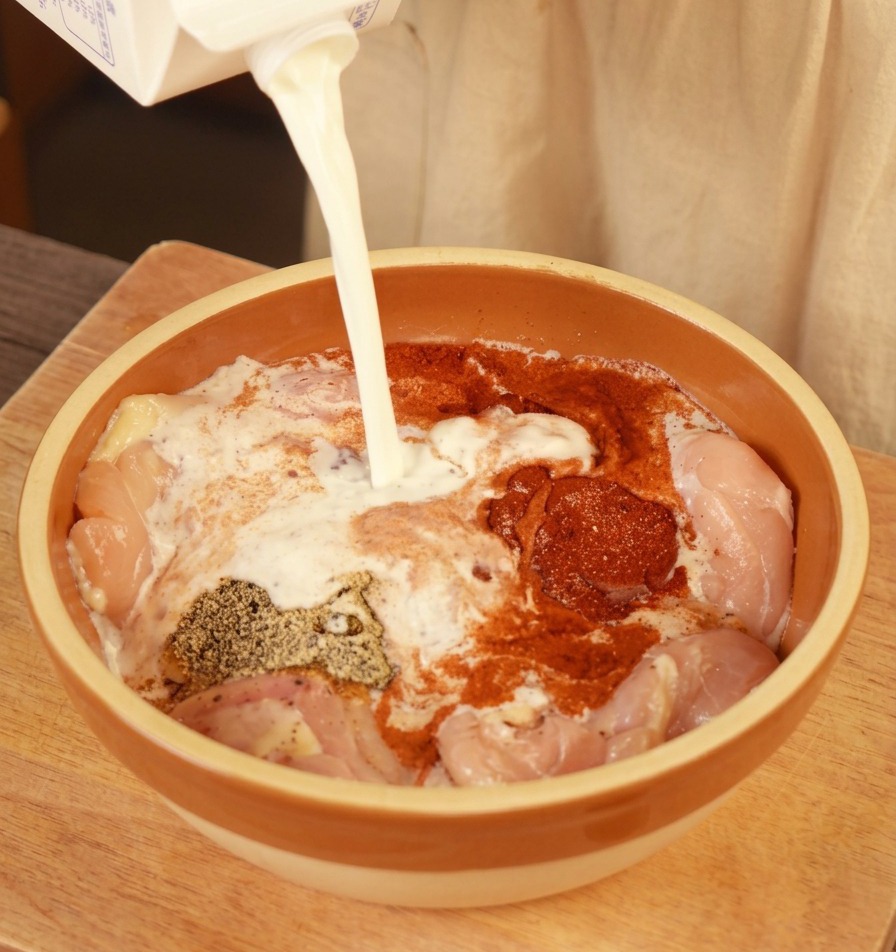 Frische Milch wird aus einem Karton in eine Schüssel mit gewürzten rohen Hähnchenoberschenkeln gegossen.