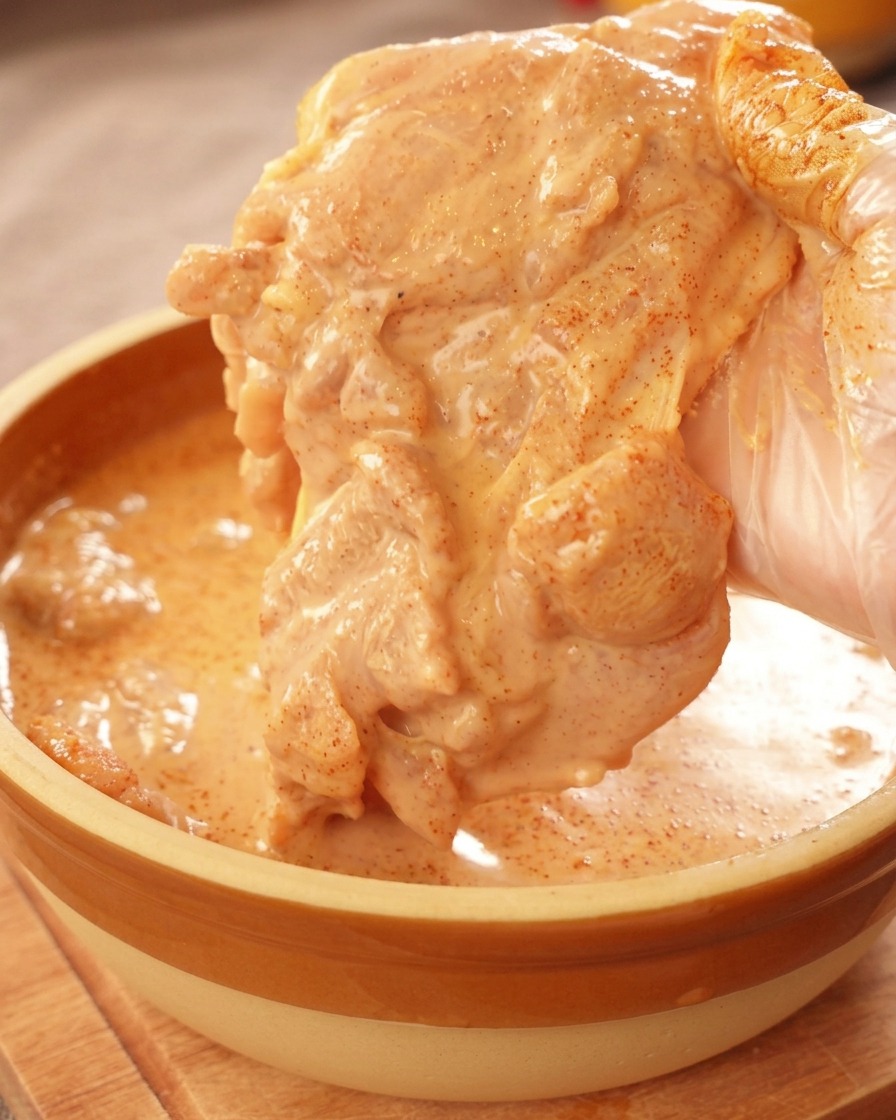Eine behandschuhte Hand hebt ein Stück Hähnchenoberschenkel an, das großzügig mit einer dicken, orangefarbenen Marinade bedeckt ist.
