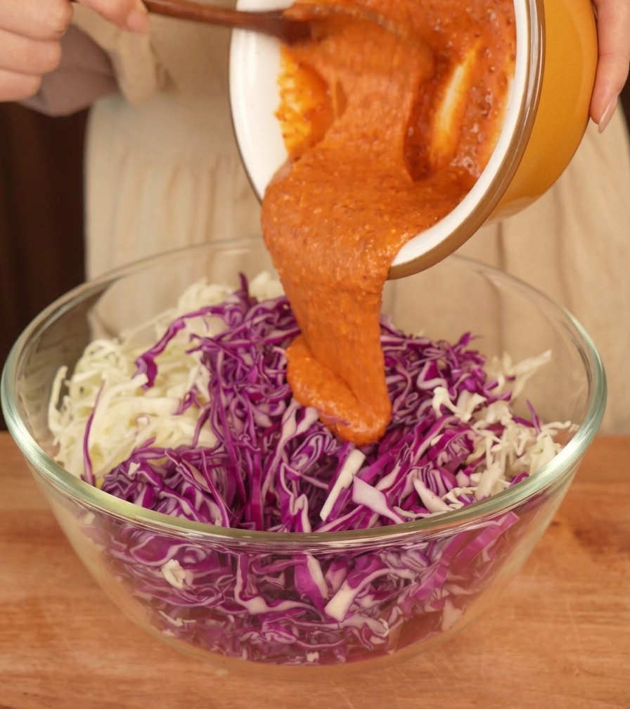 Cremiges orangefarbenes Dressing wird aus einer gelben Keramikschüssel über einen Berg aus geraspeltem Rot- und Weißkohl in einer Glasschüssel gegossen.