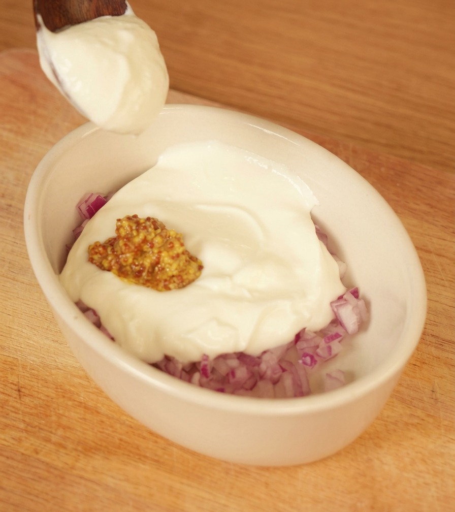 Ein Löffel dicker weißer Joghurt wird in eine kleine weiße Schüssel mit gewürfelten roten Zwiebeln und Senf gegeben.