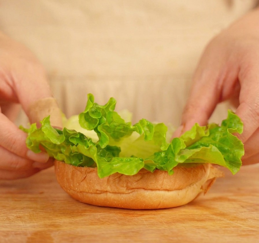 Hände platzieren vorsichtig grüne Salatblätter auf die Unterseite eines getoasteten Burgerbrötchens.