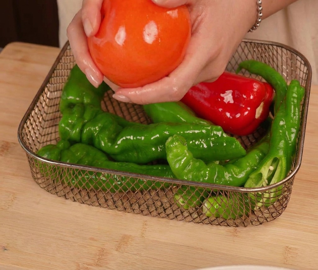 Hände legen eine glänzende, eingeölte Tomate neben mehrere grüne und rote Chilis in den Korb einer Heißluftfritteuse.