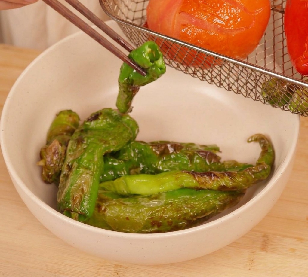 Essstäbchen heben eine stark geröstete grüne Chilischote aus dem Korb einer Heißluftfritteuse, in dem sich eine geröstete Tomate und weitere Chilis befinden.