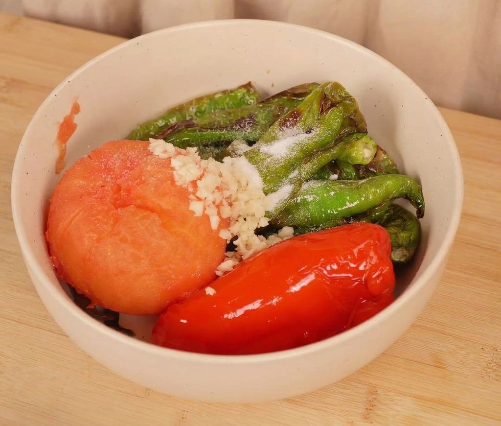 Eine Schüssel mit gerösteten Chilis und einer Tomate, belegt mit frischem gehacktem Knoblauch und einer Prise Salz, kurz vor dem Vermengen.