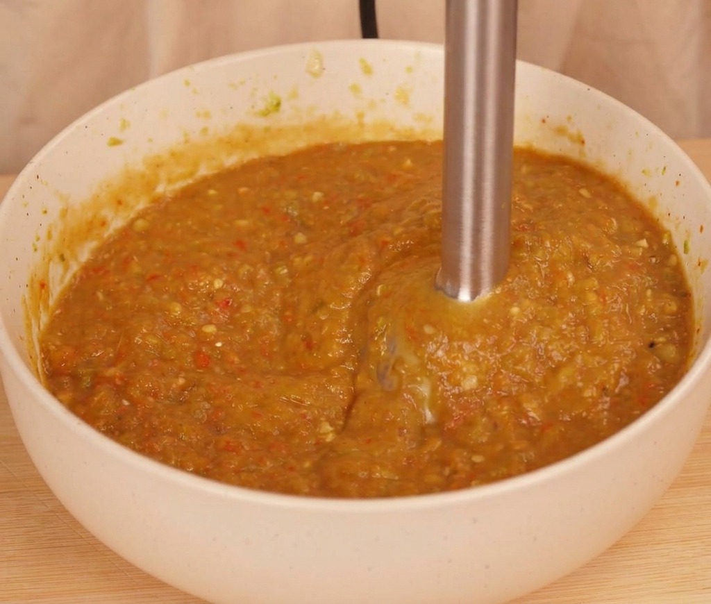 Ein Pürierstab verarbeitet geröstete Chilis und Tomate in einer weißen Schüssel zu einer dickflüssigen, rustikalen orange-braunen Chili-Sauce.