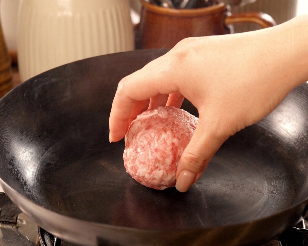 Eine Hand legt eine geformte Kugel rohes Rinderhackfleisch in einen rauchend heißen, ungefetteten gusseisernen Wok.