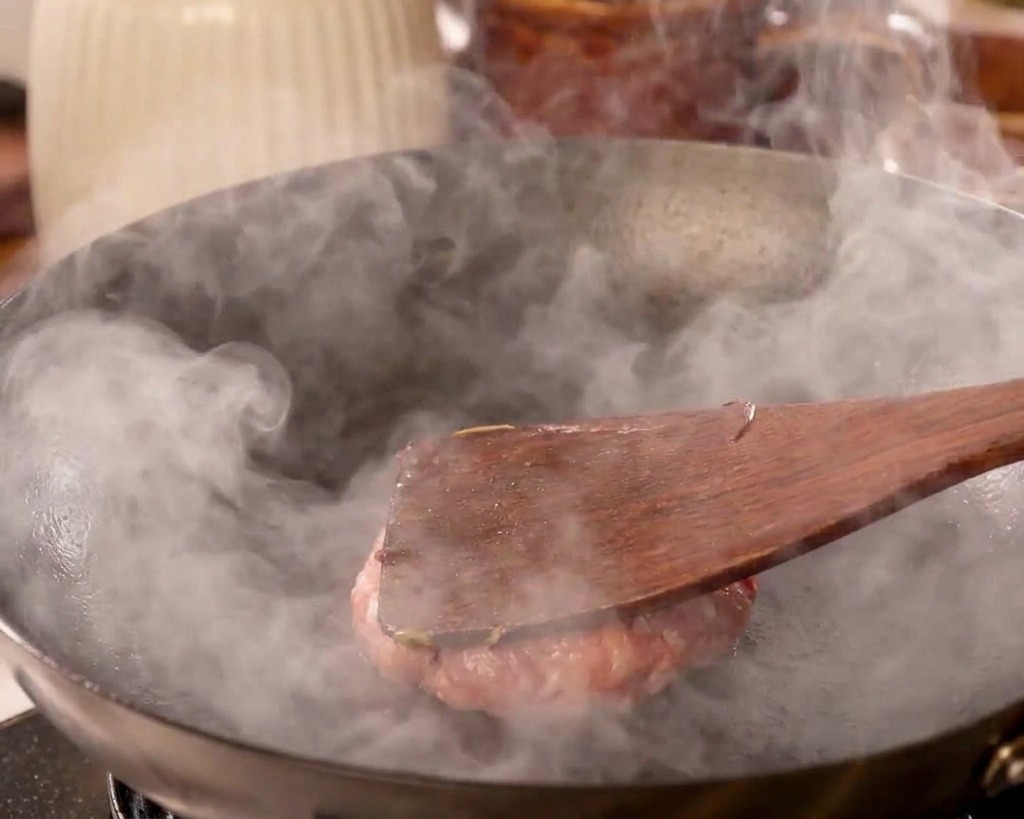 Ein flacher Holzspatel drückt ein Rindfleisch-Patty fest nach unten, flach auf die Oberfläche eines heißen, rauchenden Woks.