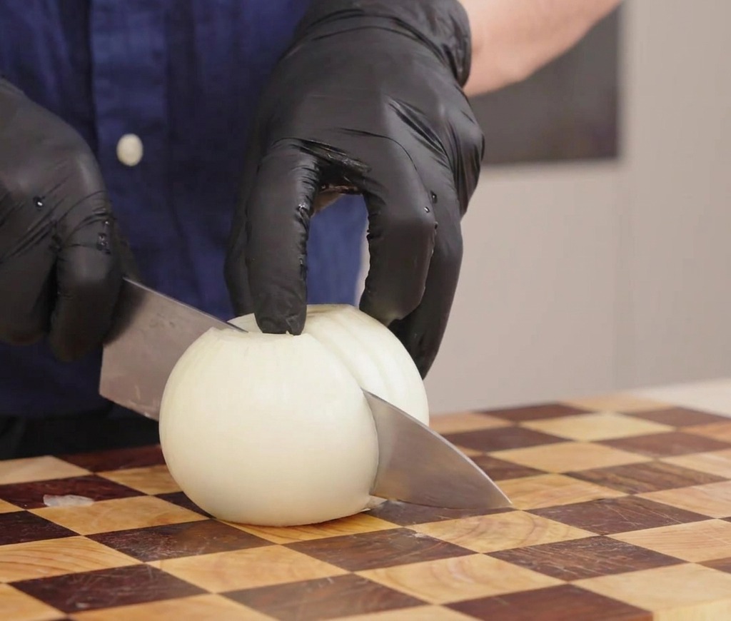 Mit Handschuhen bekleidete Hände schneiden eine weiße Zwiebel auf einem karierten Holzschneidebrett in Streifen.