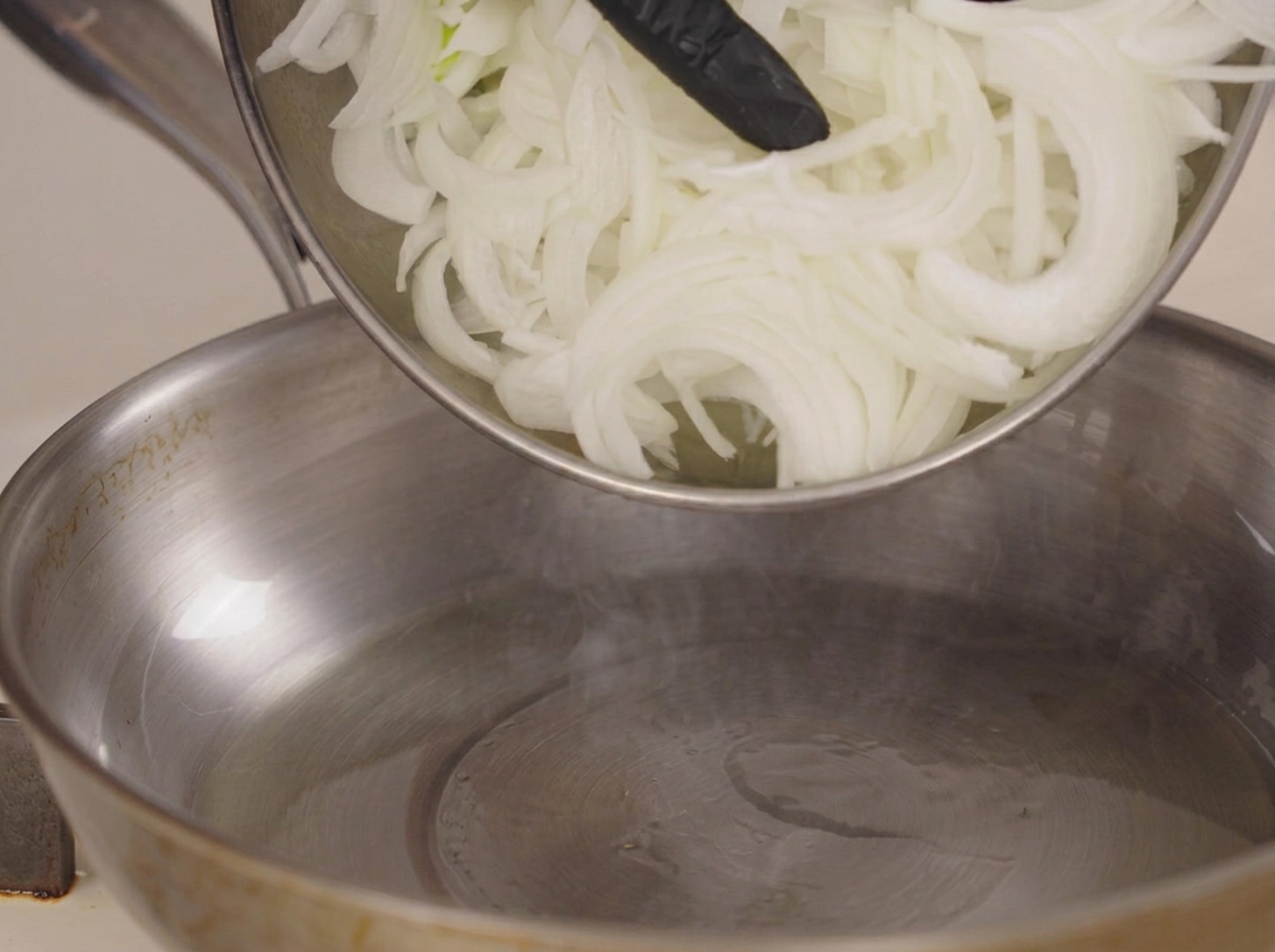 Rohe Zwiebelstreifen werden in eine Edelstahlpfanne gegeben.
