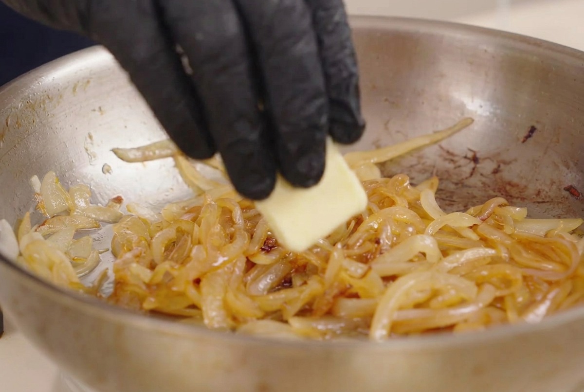 Eine Hand gibt ein Stück Butter in eine Edelstahlpfanne mit angedünsteten weißen Zwiebeln und dunkler Würzsoße.