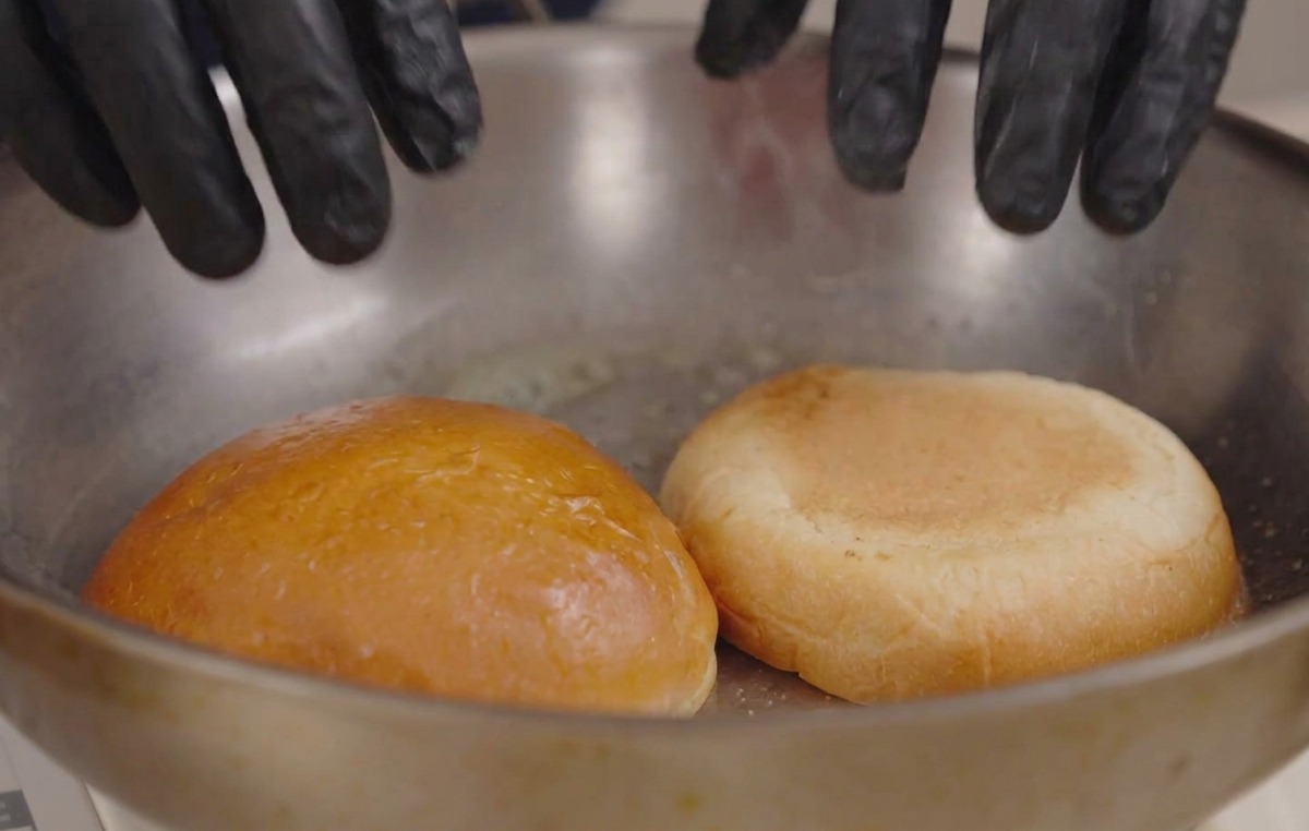 Hände in schwarzen Handschuhen drücken zwei Brötchenhälften mit der Schnittfläche nach unten in eine heiße Pfanne mit geschmolzener Butter.