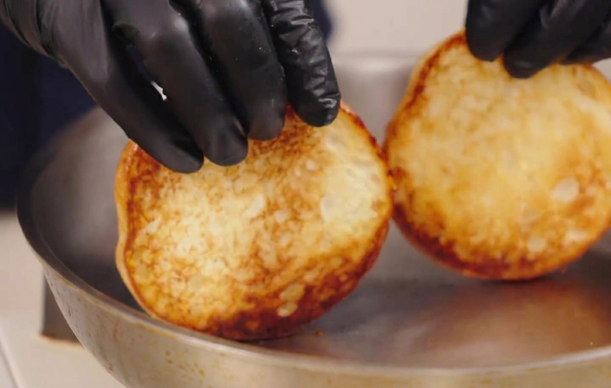 Behandschuhte Hände heben zwei geröstete Burgerbrötchen aus einer Pfanne und zeigen die perfekt gebräunte, karamellisierte Unterseite.